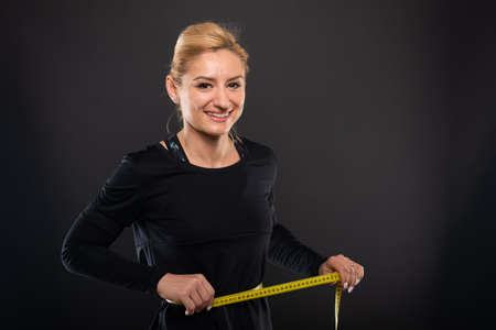 Pretty gym female trainer holding measuring herself and smiling on black background with copypsace...