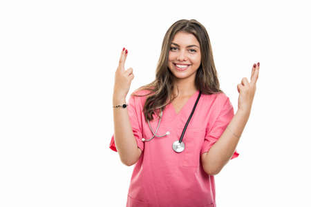 Portrait of beautiful young doctor wearing scrubs showing double  fingers crossed gesture isolated on white background with copy space advertising areaの写真素材
