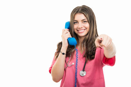 Portrait of beautiful young doctor talking at telephone pointing camera isolated on white background with copy space advertising areaの写真素材