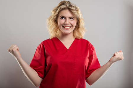 Portrait of female nurse wearing red scrub making winner gesture on studio gray background with copy space advertising areaの写真素材