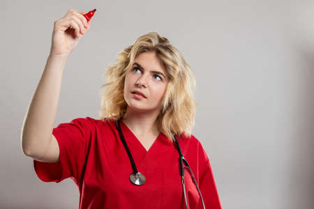 Portrait of female nurse wearing red scrub writing with marker on studio gray background with copy space advertising areaの写真素材