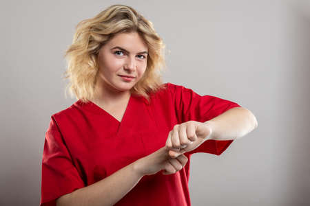 Portrait of female nurse wearing red scrub making late gesture on studio gray background with copy space advertising areaの写真素材