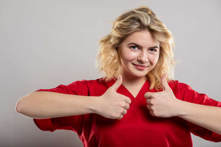 Portrait of female nurse wearing red scrub showing double thumb up on studio gray background with copy space advertising areaの写真素材