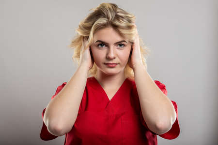 Portrait of nurse wearing red scrub covering ears like deaf concept on studio gray background