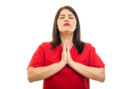 Portrait of beautiful young doctor wearing scrubs showing prayer gesture isolated on white...