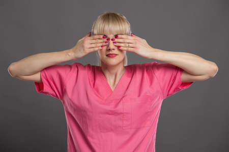 Portrait of young attractive female nurse covering eyes like blind gesture on gray backgroundの写真素材