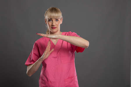 Portrait of young attractive female nurse showing time out gesture on gray background with copy...