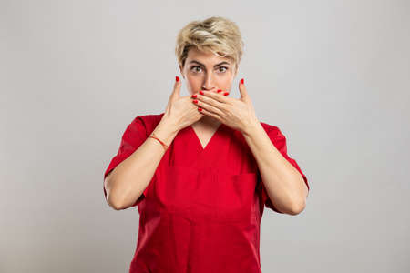 Portrait of young attractive female nurse covering mouth like mute gesture on grey backgroundの写真素材