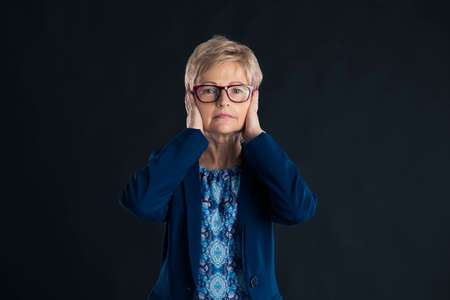 Senior businesswoman having her hands up to her ears so she does not hear on a dark backgroundの写真素材