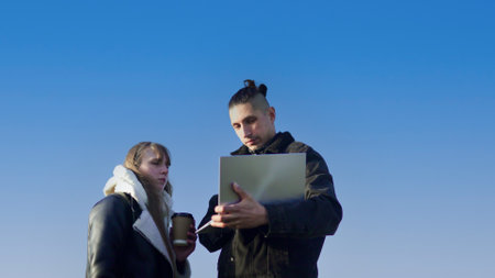 Two young professionals collaborating outdoors, using laptop and drinking coffee under blue sky