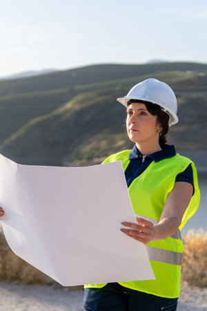 female engineer wearing a fluorescent safety vest examining a blueprint at a construction site with...