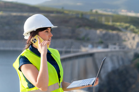 Female engineer managing infrastructure, talking on phone and using laptop near damの写真素材