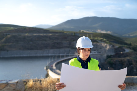 Female engineer examining blueprint by a dam reservoir in mountainous landscape