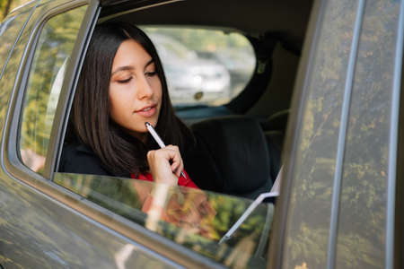 Stylish woman touching pen to her chin in car. Woman touching her chin with pen while sitting on...