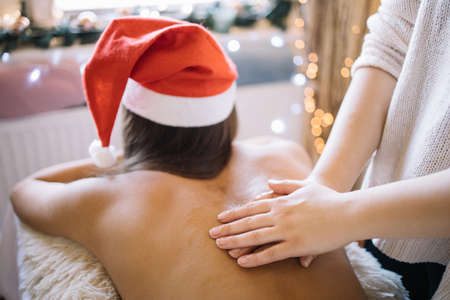 Masseuse massaging female body in saloon with lights. Woman wearing Santa hat and lying on massage table while having massage with oil in SPA center decorated with Christmas lights, with copyspace. Image can be used in websites, advertising, blogs and articles.の写真素材