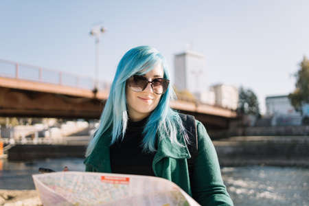 Back view of woman taking a photo of river. Backside of woman with blue hair wearing backpack and...