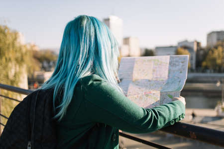 Lost solo tourist girl looking for location into a map. Back of tourist woman with blue hair on vacation searching for direction on paper map outdoors with copyspace. Image can be used in websites, advertising, blogs and articlesの写真素材
