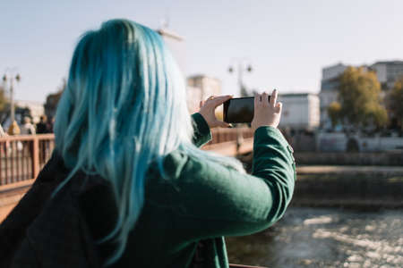 Back of out of focus woman taking picture. Backside of girl with blue hair making photo with her phone of river while standing near bridge, with copyspace. Ideal for blogs, websites and magazines.の写真素材