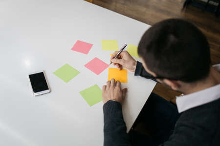 Top view of sticky notes and phone on desk. Office worker holding pen and writing on blank colorful...