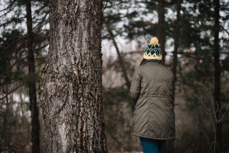 Back view of a girl standing next to tree. Back of a woman with winter clothes stands next to old tree in the forest.の写真素材