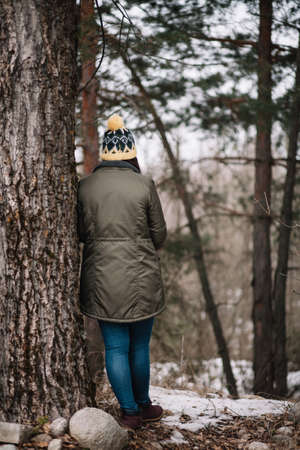 Back of a girl leaning on big tree. Back view of a girl wearing jacket and knitted hat leaning against old tree during winter.の写真素材