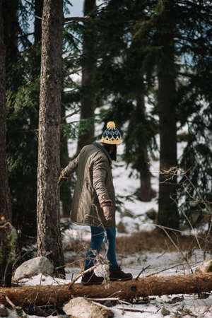 Young girl trying to balance on fallen tree. Lady wearing hat, jacket and jeans stepping on a fallen tree in the winter snowy forest.の写真素材