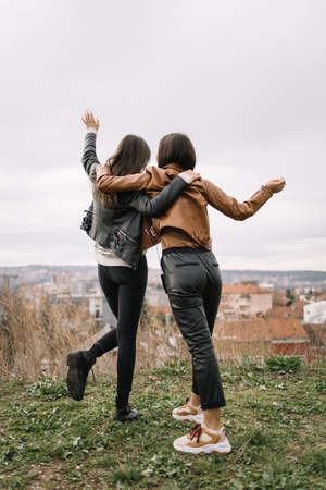 Back of girls hugging and waving with hands. View from back of fashionable women hugging while waving and looking at city.の写真素材