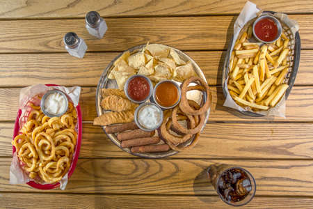 Top view of assorted snacks on wooden table. Wooden table with coke, salt and pepper, fried onion rings, fish fingers, breaded squid, chips, potatoes, sauces and tortilla.の写真素材