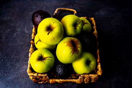 Apples and avocados in wicker platter on black surface