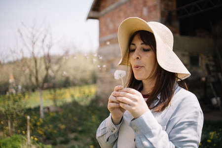 Pretty girl wearing hat blowing dandelion in backyardの写真素材