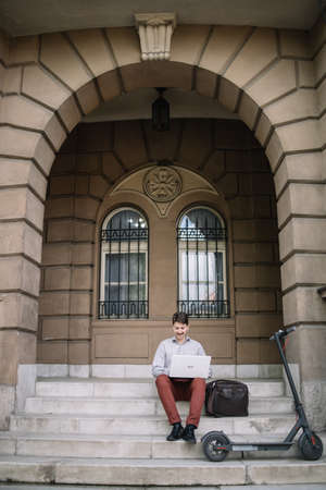 Portrait of man sitting under arch with laptop and electric scooterの写真素材