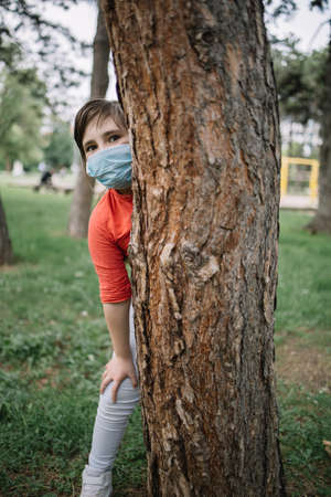 Portrait of child with medical mask playing in park. Girl wearing antivirus mask standing behind tree in city garden during coronavirus pandemic.の写真素材