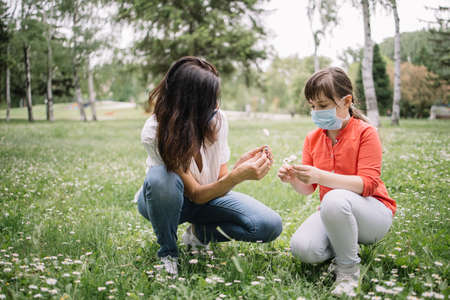 Girls with antivirus masks sitting on lawn in park and gathering flowers. Woman and kid with protection masks squatting on meadow with camomile during pandemic.の写真素材