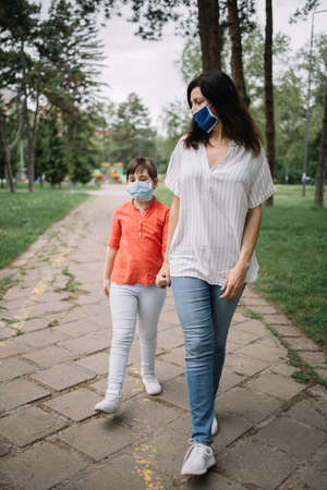 Mom and child walking on park alley while wearing antivirus masks. Woman holding little girls hand while walking in park and wearing medical masks against coronavirus.の写真素材