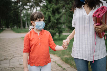 Portrait of child wearing mask and looking sad in park. Daughter and mother holding their hands while walking in nature during coronavirus pandemic.の写真素材