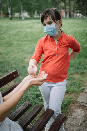 Child applying hand sanitizer to clean and disinfect female hands. Cute girl with medical mask disinfecting mothers hands with antiseptic spray while standing in park.の写真素材
