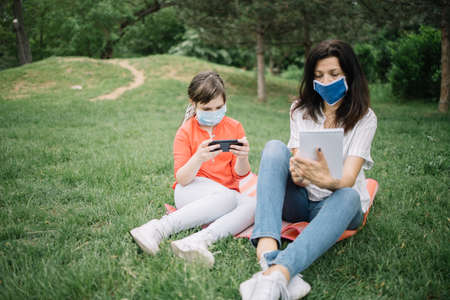Woman wearing antivirus mask while resting in park with her daughter. Little girl with antivirus mask using phone while lying on mat in forest with her mother who is holding notebook.の写真素材
