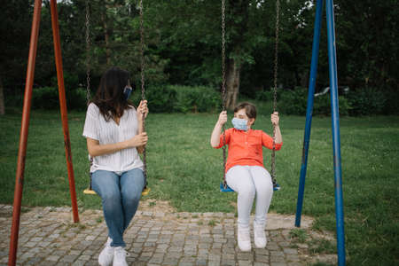 Woman and little girl on swing while wearing antivirus masks. Mother and daughter with protection masks are swinging in park.の写真素材