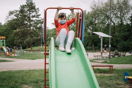 Front view of young girl with protection mask on her face going down a slide. Happy daughter preparing to free her hands from the handle to go down the slide. Other playground facilities in the back.の写真素材
