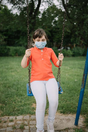 Girl with antivirus mask swinging in park during coronavirus pandemic. Cute girl child using swing...