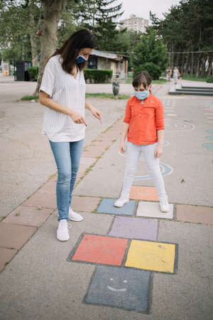 Nice woman and girl at the playground during coronavirus pandemic. Careful mother indicating her daughter how to jump on playground markings and wearing protection masksの写真素材