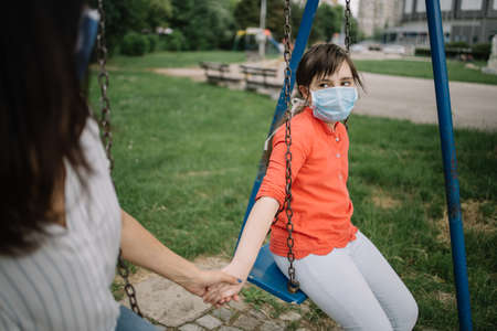 Little girl with mask holding womans hand while using swing. Portrait of girl with antivirus mask swinging in the park and holding her mothers hand.の写真素材