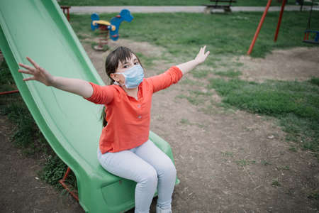 Beautiful girl with face mask sitting at bottom of the slide with hands spread. Child looking at...