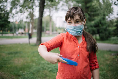 Beautiful girl with medical mask throwing frisbee. Child playing frisbee in the park after loosening of the coronavirus restrictionsの写真素材