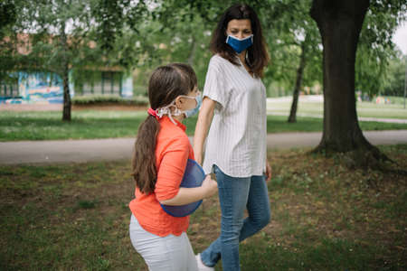 Mother and daughter with medical masks walking in the park. Brunette woman and girl protected by masks going to play frisbee outdoor.の写真素材