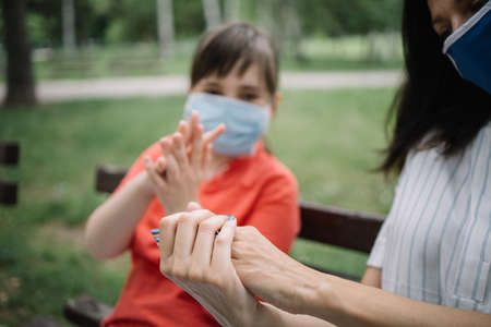 Cropped girls rubbing their hands with disinfectant while sitting on bench. Portrait of woman and child wearing antivirus masks while using hand sanitizer in park.の写真素材
