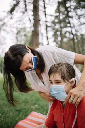 Close-up view of mother and her child wearing face masks outdoor. Child resting on ground in nature while mother is putting face mask during pandemic situation.の写真素材