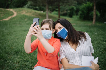 Mother and girltaking selfie in nature. Happy girls wearing antivirus mask while taking photo of themselves while resting in nature.の写真素材