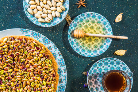 Top view of table with oriental dessert with pistachios and honey. Arabic sweet dessert of kadayif and pistachios on table with honey, wooden spoon and cup of tea.の写真素材