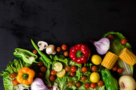 Various fresh vegetables arranged on black surface. Top view of pepper, corn, cherry tomatoes, mushrooms, lemon, onion and lettuce arranged on black table.の写真素材